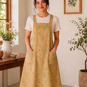 May include: A mustard yellow apron featuring a repeating pattern of small white and green wildflowers. The apron has a square neckline, thick shoulder straps, and two large square patch pockets on the front, worn over a plain white short-sleeved shirt.