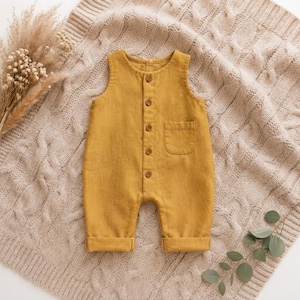 May include: A sleeveless mustard yellow romper featuring a round neckline, a front button placket with five small brown buttons, and a single patch pocket on the chest. The garment has cuffed leg openings and a simple, straight-leg silhouette.