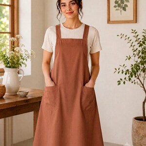 May include: A solid rust-colored apron featuring a square neckline, wide shoulder straps, and two large patch pockets on the front. The apron has a simple, clean design with a straight hemline that falls below the knees, providing full coverage.