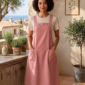 May include: A dusty rose pinafore dress featuring a square neckline, thick shoulder straps, and two large patch pockets on the front. The dress is worn over a short-sleeved, cream-colored ribbed knit top with a round neckline and a fitted silhouette.