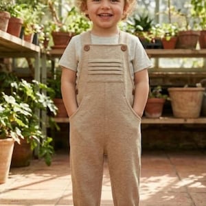 Puede incluir: Un niño con un mono marrón claro con camiseta y zapatillas a juego. El mono tiene botones de madera grandes y bolsillos. El niño sonríe en un invernadero con plantas y macetas de terracota.