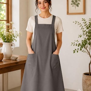 May include: A charcoal gray apron featuring a square neckline, wide shoulder straps, and two large patch pockets on the front. The apron is worn over a short-sleeved, cream-colored ribbed t-shirt, creating a layered look with a simple, clean silhouette.