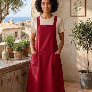 May include: A deep red apron featuring a square neckline, thick shoulder straps, and two large front patch pockets. The apron is worn over a short-sleeved, cream-colored ribbed shirt, creating a layered look with a simple, functional design.