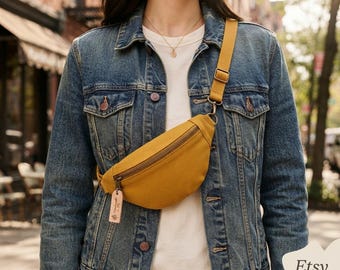May include: A mustard yellow canvas fanny pack with a top zipper closure and an adjustable strap. A small, rectangular, light-colored wooden tag hangs from the zipper pull. The bag is worn across the chest over a white shirt and a blue denim jacket.