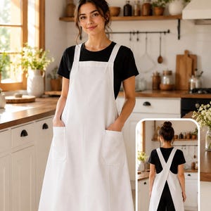 May include: A solid white apron featuring a square neckline, thick shoulder straps, and two large front patch pockets. The design includes a full-length skirt and a cross-back strap configuration, shown from both the front and back perspectives.