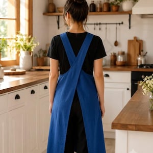 May include: A solid blue apron featuring a cross-back design with wide shoulder straps that overlap at the back. The apron has a straight hemline that falls below the waist, providing full coverage over a black short-sleeved shirt and black pants.