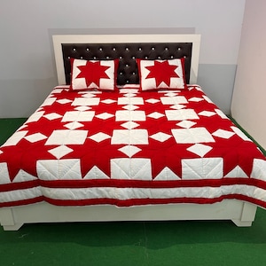 Könnte beinhalten: Ein Bett mit weißem Rahmen und einem dunkelbraunen, gesteppten Kopfteil mit silbernen Akzenten. Das Bett ist mit einer Steppdecke und zwei passenden Kissenbezügen bezogen, die ein geometrisches Muster aus roten Sternen auf weißem Grund zeigen.