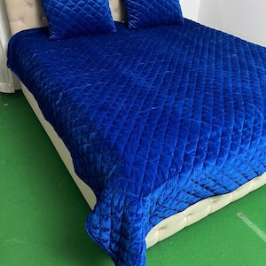 Puede incluir: Una colcha de terciopelo azul real vibrante y dos cojines cuadrados a juego, todos con un patrón acolchado de diamantes. La colcha tiene una pequeña borla en la esquina y está colocada sobre una estructura de cama beige capitoné.