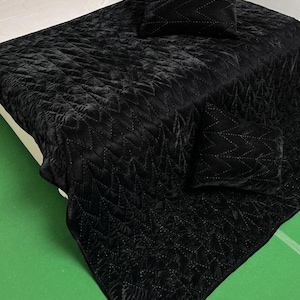 May include: A black velvet quilt set with two matching cushions. The quilt and cushions feature a stitched zigzag pattern. The bedding is displayed on a bed with a cream-coloured headboard and white sheets. The background is a green surface.