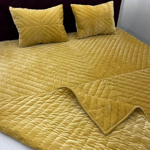May include: A bed featuring a mustard yellow quilted bedspread and two matching square pillows. The pillows have a geometric stitched pattern. The headboard is a deep burgundy colour. The bedspread is folded over at the foot of the bed.