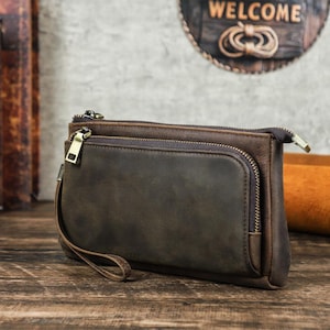 Peut inclure: Une pochette en cuir marron avec une dragonne. La pochette comporte deux compartiments zippés et une poche avant. Le cuir a une finition vieillie et les fermetures éclair sont dorées. La pochette est posée sur une surface en bois.