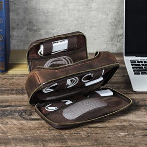 May include: Open brown leather tech organizer with multiple compartments, holding white charging cables, a USB hub, and a computer mouse. The zippered case is on a wooden surface, next to a laptop.