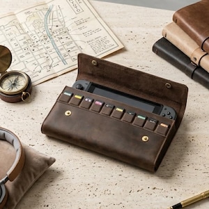 May include: A dark brown leather carrying case for a handheld gaming console, featuring a flap with two brass snap buttons and a row of eight small interior slots for game cartridges. The case is open, revealing the console screen and buttons.