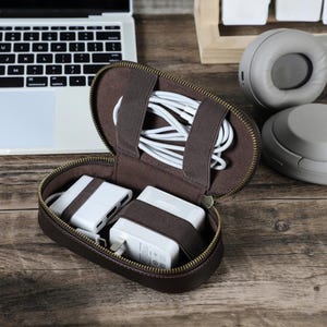 Könnte beinhalten: Eine braune Leder-Reisetasche mit Reißverschluss, in der sich weiße Ladekabel und Netzadapter befinden. Die Tasche hat braune elastische Bänder zur Sicherung des Inhalts. Ein Laptop und Kopfhörer befinden sich im Hintergrund.