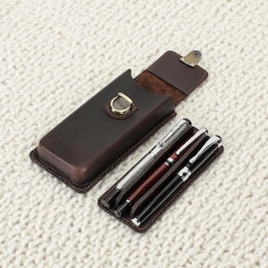 Op de afbeelding: Een bruin leren etui met een goudkleurige sluiting, open om drie pennen te laten zien. De pennen zijn zilver, bordeauxrood en zwart, elk met zilveren details. De etui ligt op een witte, getextureerde ondergrond.