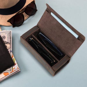 Puede incluir: Un estuche de gafas de cuero marrón abierto, que contiene dos pares de gafas. Un sombrero de paja y gafas de sol son visibles en el fondo. El estuche tiene dos compartimentos y un paño de limpieza azul claro.