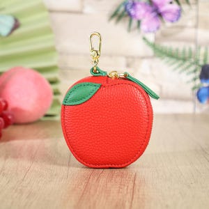 May include: A round, red, textured coin purse shaped like an apple, featuring a green leather leaf accent on the top, a gold-toned metal zip closure, and a matching gold-toned lobster clasp attached to the top for easy carrying or hanging.