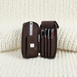 May include: A brown leather pen case, open to reveal pens, a ruler, eraser, and paper clips. The case is zippered and has individual slots for pens. The pens are black, silver, and red. The ruler is silver and marked with centimetres.