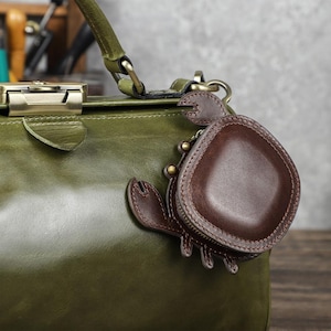 Könnte beinhalten: Eine dunkelbraune Leder-Münzbörse in Krabbenform mit Reißverschluss und Metallbeschlägen, befestigt an einer großen olivgrünen Lederhandtasche mit Metallverschluss und stabilem Griff. Der Krabbenanhänger weist sichtbare Nähte auf.