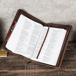 May include: Open Bible in a brown leather case with a red pen. The Bible is open, revealing text, and has a brown bookmark. The case has a zip closure.