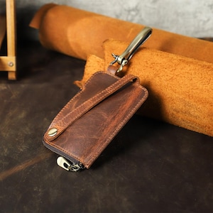 May include: A brown leather key holder with a zip and metal clasp. The key holder has a distressed finish and a strap. The background includes a roll of brown leather and a dark brown surface.