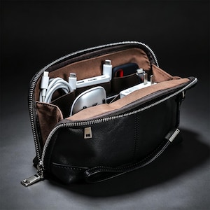 May include: A black leather zipped pouch with a brown interior lining, featuring multiple internal compartments organised with various electronic accessories, including white charging cables, a power bank, and a small white computer mouse.