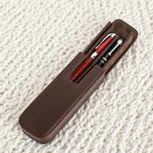 May include: A brown leather pen case containing a red pen and a black pen. The case is rectangular with a flap and a soft interior. The pens have silver detailing. The case is on a white, textured surface.