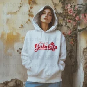 Può includere: Una felpa con cappuccio bianca con il nome "Gabriela" in rosso sul petto. La felpa ha un cappuccio con coulisse e una tasca frontale. La modella indossa jeans blu.