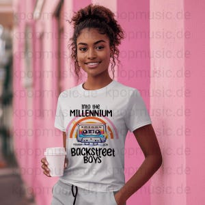May include: White t-shirt featuring a colorful graphic of a cassette tape with a rainbow and the text "Into the Millennium Backstreet Boys 2026". The shirt is worn by a person holding a white cup.