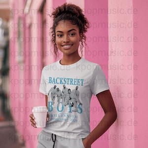 Op de afbeelding: Wit T-shirt met de tekst "BACKSTREET BOYS" in blauw en een foto van de band. Het shirt heeft ook "INTO THE MILLENNIUM Düsseldorf 2026". De persoon houdt een witte beker vast.