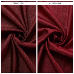 May include: Close-up of two sections of a deep burgundy fabric, with visible folds and textures. The fabric has a subtle sheen, and the color is consistent throughout. The text "CLUNY - 002" and "CLUNY - 003" are visible at the top.