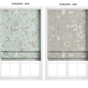 May include: Two Roman shades with floral patterns. One shade is light blue with brown and white floral designs. The other is gray with white and brown floral designs. The shades are mounted inside white window frames.