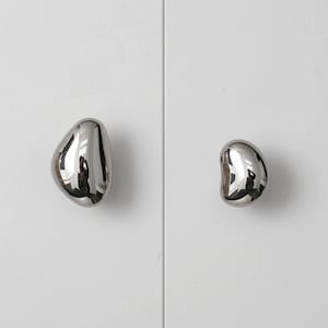 Pode incluir: Duas puxadores de armário cromados polidos com formato orgânico, semelhante a seixos, montados em uma porta de armário branca. Os puxadores estão posicionados no lado direito da porta, sendo um ligeiramente maior que o outro.