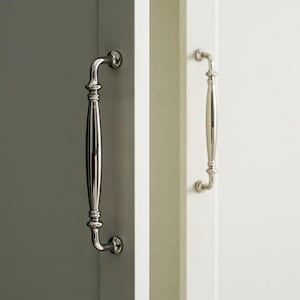 Op de afbeelding: Close-up van twee gepolijste zilveren kastgrepen. Eén handvat is bevestigd aan een grijze kastdeur, de andere aan een witte deur. De handgrepen hebben een klassiek, elegant ontwerp met een gebogen basis en een lange, cilindrische grip.