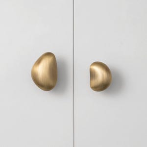 May include: Two brass cabinet pulls with an organic, pebble-like shape are mounted on a white cabinet door. The pulls have a smooth, polished finish and are positioned on either side of a vertical seam.