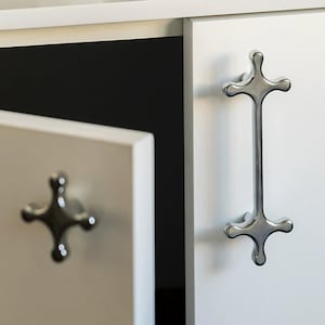 Puede incluir: Puertas de armario blancas con tiradores cromados en forma de cruz. Una puerta está entreabierta, revelando un interior oscuro. Los tiradores tienen un diseño moderno y minimalista.