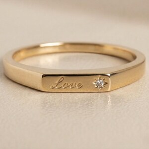 Könnte beinhalten: Ein goldfarbener Ring mit dem Wort "Love" auf der Vorderseite eingraviert. Ein kleiner, sternförmiger Diamant ist neben dem Wort gesetzt. Der Ring hat ein flaches, rechteckiges Design.