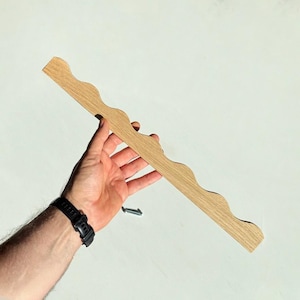 Puede incluir: Una tira larga y estrecha de madera de color claro que presenta un borde decorativo ondulado o festoneado en un lado. La pieza de madera tiene un patrón de veta natural y se muestra junto a un pequeño tornillo de metal plateado para montaje.