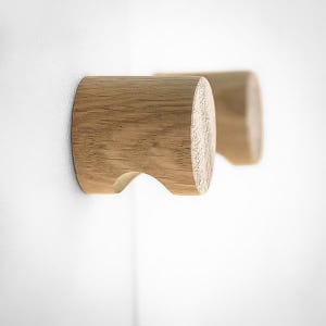 May include: Two cylindrical wall hooks crafted from light-colored natural wood with a visible grain pattern. Each hook features a smooth, rounded front face and a curved cutout on the underside, designed for mounting flush against a flat surface.