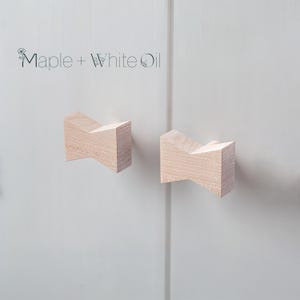 May include: Two light-colored wooden cabinet knobs with a bow-tie shape attached to a white cabinet door. The text Maple + White Oil is printed in black above the knobs, accompanied by a small line drawing of a flower.
