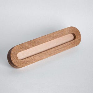Puede incluir: Un objeto de madera de forma ovalada alargada con un acabado liso de color marrón claro. La pieza presenta un canal horizontal empotrado que recorre el centro, revelando una textura de veta de madera natural más clara en la cavidad interior.