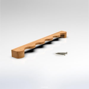 Puede incluir: Un tirador de mueble de madera de color marrón claro con un borde festoneado que presenta una serie de curvas redondeadas en la parte superior. El tirador incluye dos tornillos de metal plateado para su instalación y veta natural.