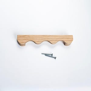 May include: A wooden wall-mounted shelf with a wavy design, featuring a natural wood grain finish. Two silver screws are positioned below the shelf. The shelf is designed for decorative or storage purposes.