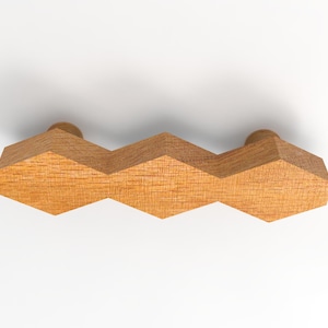 Könnte beinhalten: Ein hellbrauner Möbelgriff aus Holz mit einem geometrischen Design aus drei verbundenen sechseckigen Formen. Der Griff weist eine sichtbare Holzmaserung und zwei zylindrische Befestigungspunkte auf der Rückseite auf.