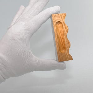 May include: A rectangular wooden object with a wavy edge and a carved-out groove. The wood has a light brown color with visible grain patterns. The object is held in a white gloved hand.