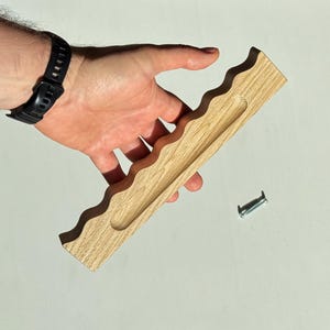 Puede incluir: Un tirador de madera de color claro con un diseño de borde ondulado y festoneado en un lado y una ranura alargada y empotrada en el centro. El tirador viene acompañado de un tornillo metálico de color plateado para su instalación.
