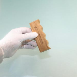 Pode incluir: Um cabo de madeira castanho-claro com um acabamento de grão natural e liso. O objeto apresenta uma forma retangular com uma borda ondulada e contornada de um lado e uma ranhura vertical longa, estreita e rebaixada no centro.