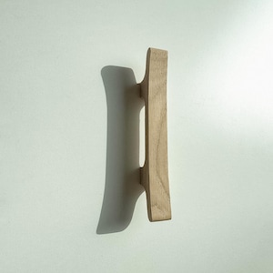 Peut inclure: Une poignée de placard verticale en bois brun clair avec une forme allongée et incurvée, présentant une texture de grain de bois naturel visible. La poignée possède deux points de fixation sur une surface unie et claire.