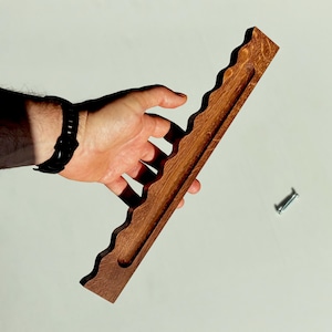 Op de afbeelding: Een lange, rechthoekige bruine houten plank met een golvende, geschulpte rand aan één kant en een verzonken groef die door het midden loopt. Een enkele zilveren metalen schroef is apart aan de zijkant van het houten stuk geplaatst.