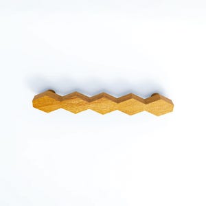 May include: A horizontal cabinet pull handle crafted from light brown wood, featuring a repeating geometric pattern of five connected hexagons. The natural wood grain is visible across the surface of the elongated, multi-faceted hardware piece.